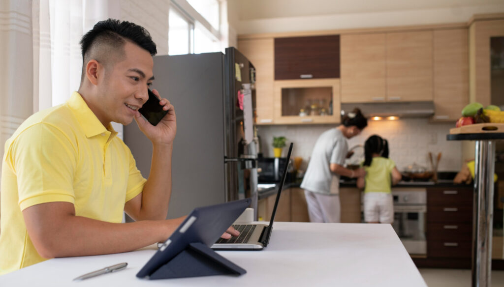 man working from home in a modern kitchen while his family prepares a meal in the background